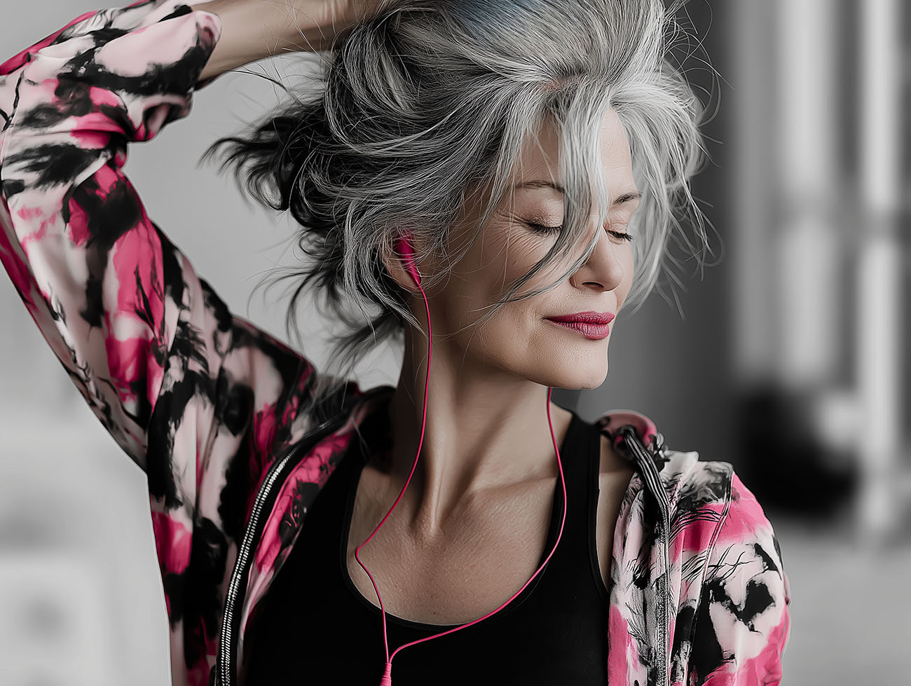 Frau mit grauen Haaren hört entspannt Musik über pinke Kopfhörer, trägt eine pink-schwarze Jacke und lächelt mit geschlossenen Augen.
