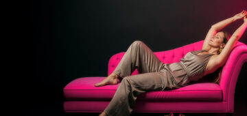 Frau liegt entspannt auf einer leuchtend pinken Chaiselongue vor dunklem Hintergrund; elegante Pose, beige Jumpsuit, neonpinke High Heels am Boden – moderne ästhetische Titelgrafik für einen Branding-Artikel über multisensorische Markenidentität.