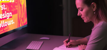 Frau sitzt an einem Schreibtisch und schreibt in ein Notizbuch, während auf einem Computerbildschirm ein farbintensives Branding-Motiv mit dem Text ‚Strategy first. Design follows.‘ angezeigt wird; Szene mit pink-violettem Licht.