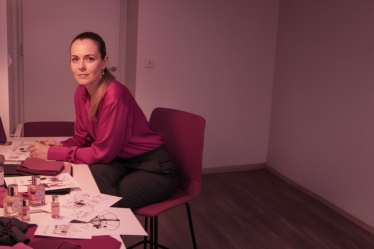 Frau sitzt an einem Tisch und arbeitet mit Duftfläschchen, Textilien und Skizzen; kreativer Arbeitsplatz in warmem, pink-violettem Licht – visuelle Darstellung eines Airdesign- und Branding-Prozesses.