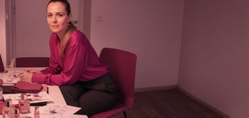 Frau sitzt an einem Tisch und arbeitet mit Duftfläschchen, Textilien und Skizzen; kreativer Arbeitsplatz in warmem, pink-violettem Licht – visuelle Darstellung eines Airdesign- und Branding-Prozesses.